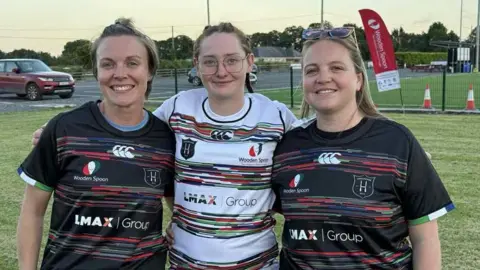 Ellie Ferrie Three women, one in a white rugby shirt and two in black rugby shirts, standing on a rugby pitch with their arms around each other and smiling at the camera.