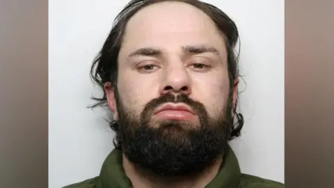 A custody photograph showing a person with dark hair and a beard against a plain light-coloured background.