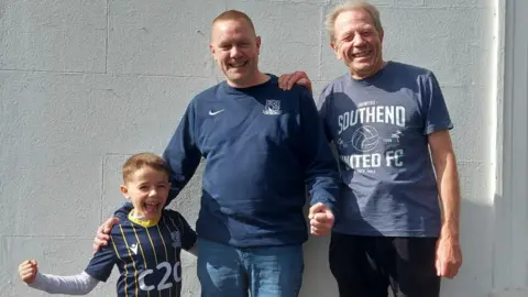 A boy with a Southend United shirt, a man with a Southend United jumper, and an older man with a Southend United T-shirt. All of them have their arms round each other.