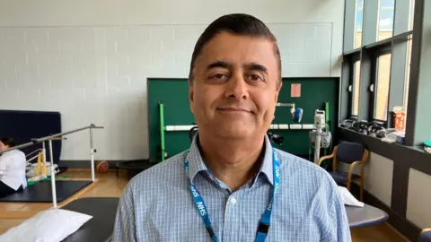 Dr Ram Hariharan is pictured in a hospital room being used for a rehabilitation session wearing a blue shirt with an NHS ID around his neck