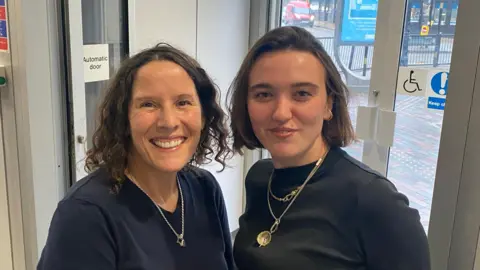 Robert Constantin/BBC Becky Rooney with curly dark hair wearing a blue top and a silver necklace. Tayla Halfacre with medium-length straight dark hair wearing a blue top and a gold neckless. There are glass doors behind them.