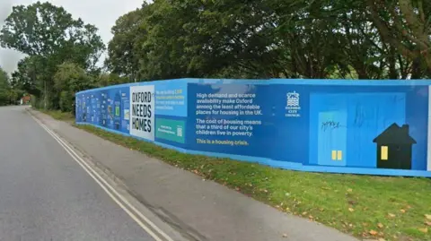 A blue hoarding erected around the chosen development site that reads Oxford Needs Homes. It is by the side of the road. The city council's logo, more information and an image of a house are also on. It is a cloudy day.