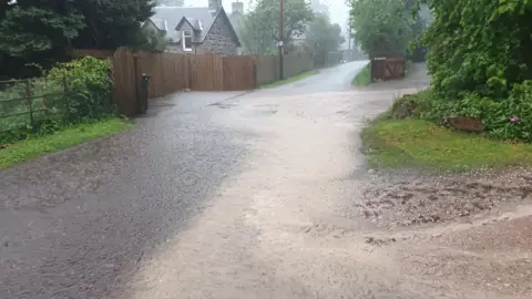 Muddy water flows down a rural road. The road is flanked by green hedges and one side of the road is a traditional, stone-built house with a peaked slate roof.