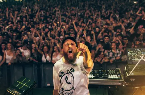 A man with a beard and black cap points at the camera as he DJS in front of a large crowd at Love Saves The Day in Bristol