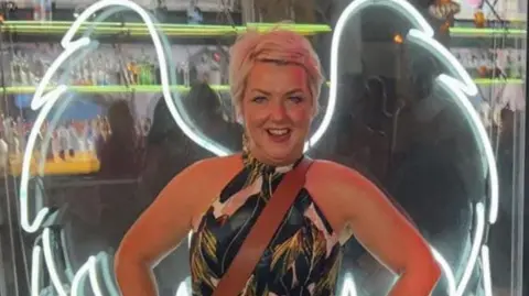 A woman with short blonde hair. She is wearing a dress and there are lights in the shape of angel wings behind her.