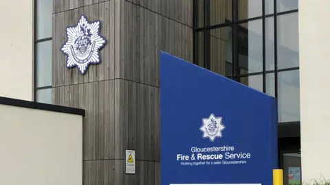 Gloucestershire Fire and Rescue Service HQ. The building has a large glass panel section and a wooden section which has the white and blue fire service logo. A blue sign can be seen to the right of the frame. It reads "Gloucestershire Fire and Rescue Service". 