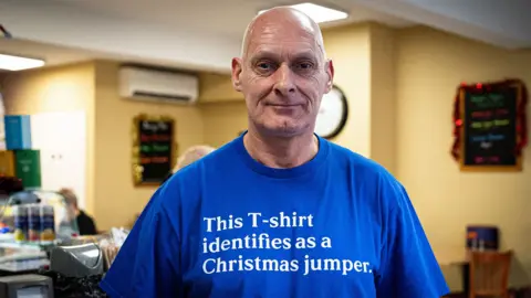 A bald man in a royal blue T-shirt bearing the words "This T-shirt identifies as a Christmas jumper" stands with his back to the counter in his coffee shop, a customer on the other side of it.