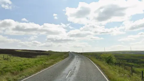 Google A road running up to the horizon with moorland on either side. Above is a blue sky with some clouds. Power lines can be seen on the horizon in the distance.
