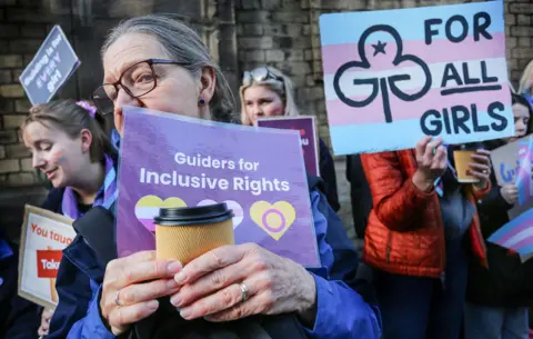 Andrea Domeniconi / LightRocket via Getty Images A person holds up a sign saying 'Guiders for inclusive rights', next to another placard in the Trans flag colours saying 'for all girls' next to a Girlguiding logo, with other demonstrators in frame, in December in London.