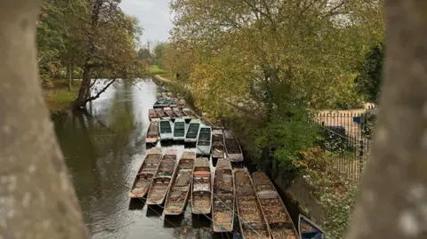 Esther J A view taken from Magdalen bridge in Oxford of punts on the River Cherwell.  Two of the bridge's stone balusters frame the edges of the picture. The punts on the water are filled with autumn leaves and are tied to the riverside. Metal railings line the waters edge.