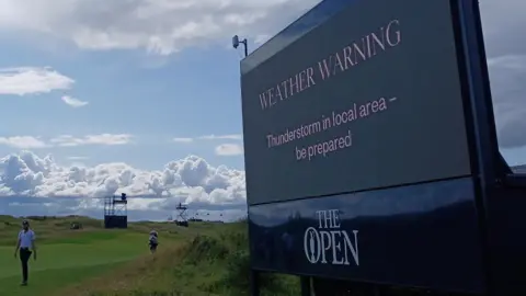 A large sign at a green at The Open saying "Weather Warning. Thunderstorm in local area - be prepared". Sun is shining with clouds approaching. People are walking along the green.