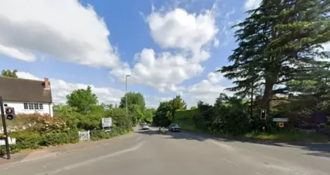 The location of the fatal collision in Ridgway Road, Farnham; the junction has both left and right turns, and traffic lights on each side. On the left hand side there is a white house, and on the right hand side there is a big tree.