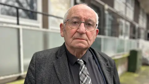Lord John Bird wearing glasses and a grey jacket over a black shirt and black and white tie. He is standing in front of a black of flats