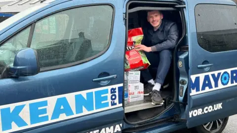 Richard Edwards/BBC Keane Duncan sits in a van, with the side door open. The van has "Keane for Mayor" written on it, while a bag of Yorkshire Tea teabags is also visible.