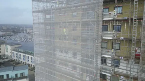 Image of building work on the outside of the apartment block. It is covered in scaffolding, with a netting outside, and man on it dressed in fluorescent clothing. A marina is visible in the background.
