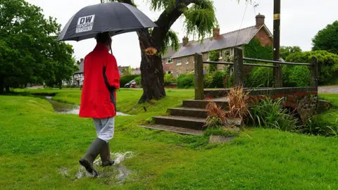 A person walks with an umbrella in a waterlogged area of grass in front of a foot bridge in the village of Charlton on 22 May