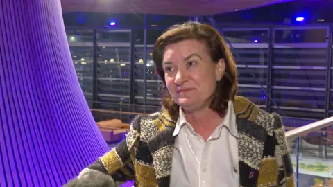 A woman with brown medium length hair looks to the right of the camera. She has a multicolored but primarily yellow jacket and a white shirt on. Behind her is a bright purple nearly cylindrical design feature and a large open plan room with light brown sofas and windows that go all the way to the top of the wall. 