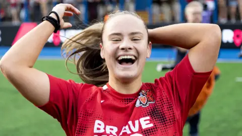 Ffion smiling on pitch in her red rugby kit. She is putting her hair in a ponytail while looking at the camera. 