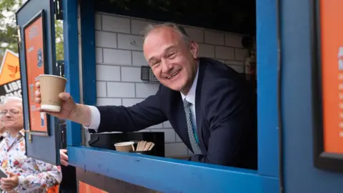 PA Media Ed Davey smiling and leaning out of a window holding a coffee.