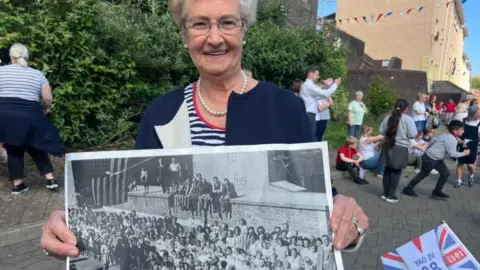 Helen Park is in a striped blue and white top and navy blazer. She is wearing glasses and pearls.
She is holding a large black and white photograph.
A number of children are in the background. 