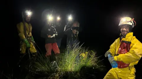 Irish Cave Rescue Organisation Five men stand on grass in the dark. They have helmets with torches on them. They are wearing waterproof overalls and carrying different tools.