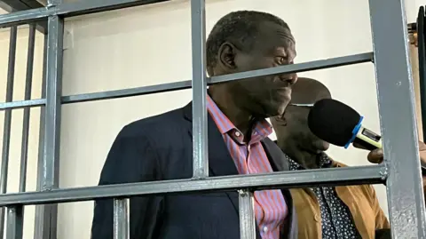 Kizza Besigye in a black blazer and striped shirt, standing behind bars at the military court in Kampala, Uganda - 20 November 2024