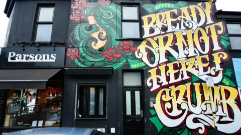Upfest A black shop has Inkie graffiti design on its wall. The design is a woman who has long wavy green hair with red flowers in it. Next to the woman is large writing which reads: 'Bready or not here I crumb'.
