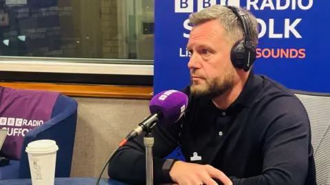 A man is sat in front of a purple microphone and wearing headphones inside a radio studio