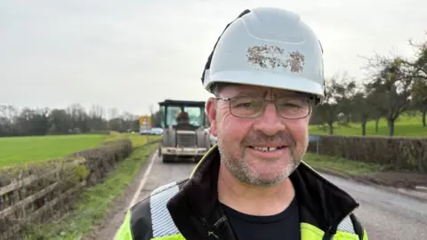 A man is wearing a white hard hat and a yellow and black high visibility coat. He is standing on a road bordered by a hedgerow and green fields. Behind him, in the distance is a large tractor.