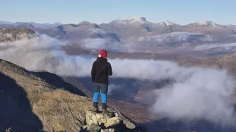 Milo Beswick Person wearing a red Santa hat standing overlooking a mist covered valley in between mountains