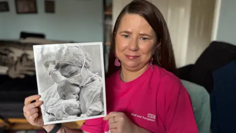 Kaylee Davidson-Olley, wearing a pink shirt which says NHS Blood and Transplant, and wearing pink heart shaped earrings holds up a black and white picture which shows her being held by her late mother, Carol. 