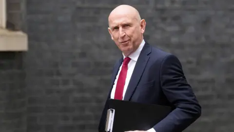 John Healey walks past No 10. He is bald and has a clipboard under his arm. He wears a navy suit and red tie.