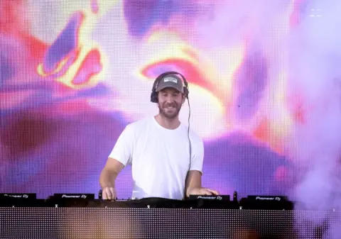 Getty Images DJ Calvin Harris in a white T-Shirt at a turntable in front of a colourful and slightly psychedelic background