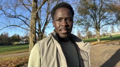 A man with curly black hair and black facial hair is wearing a grey jumper n=and beige jacket. He is standing in a park with a concrete path, grass, and brown and yellow leaves scattered on the floor