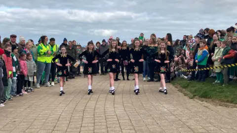 A group of five young girls perform Irish dancing in front of a crowd in Ballycastle.  The girls all have long hair and are wearing matching black dancing costumes.  They all have their hands on their hips and their right feet pointed. 