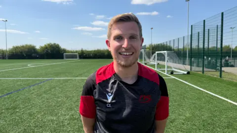 Ollie Conopo/BBC Callum in a black and red top, smiling, standing on a green artificial football pitch.