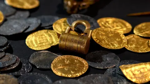 Collection of coins on table, some gold-coloured and others worn and dark with engravings and a combination lock