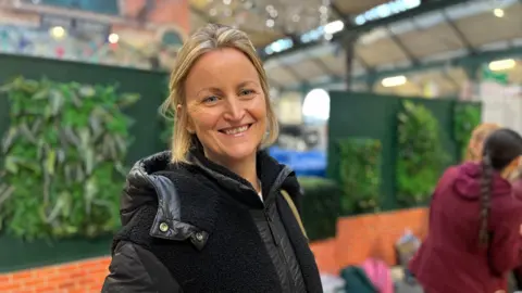 Mrs Conlon has short blonde hair and is smiling. She is wearing a black coat with a bag strap over her left shoulder. There is a red brick wall in the background with green leaves. 