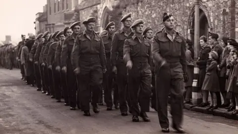 Ely Museum A black and white image showing men from the Cambridgeshire Regiment marching past Ely's war memorial. They are in Army uniform. They are being watched by men, women and children.