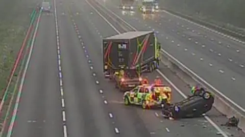 A motorway dual carriageway. A car has overturned and is on its roof and a police car and a lorry parked nearby. 