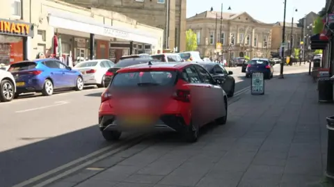 Red car parked with one side on the pavement and with the other set of wheels on double yellow lines on a high street with lots of cars in parking bays on the other side of the street. Further up the street, two more cars are stationary on the double yellow lines.