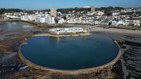 BBC The Havre des Pas lido at low tide. The pool is round and full of water and is surrounded by wet sand with lying seaweed on it. The seafront is lined with buildings.