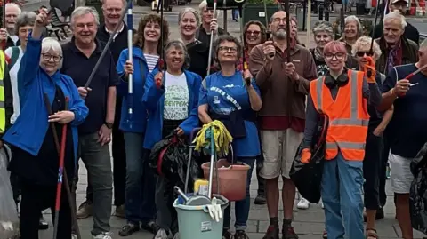 People, some in hi-vis jackets, pose for the camera in the foreground. Many are carrying cleaning equipment. Buildings are in the background.