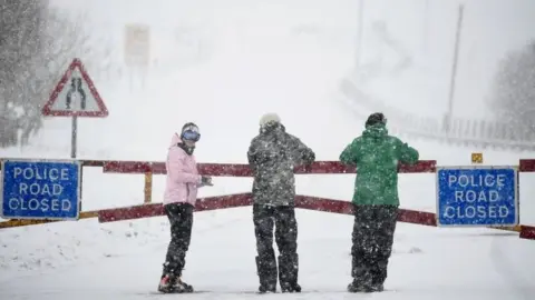 Getty Images Members of the public stand near a closed road in the snow