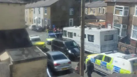 A number of police vehicles and officers stand behind a cordon in a street of terraced houses.
