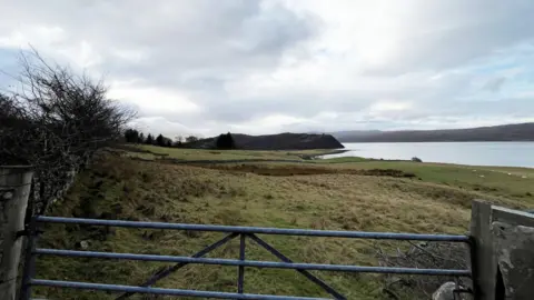 A field and body of water in Tongue, with a gate in the foreground.