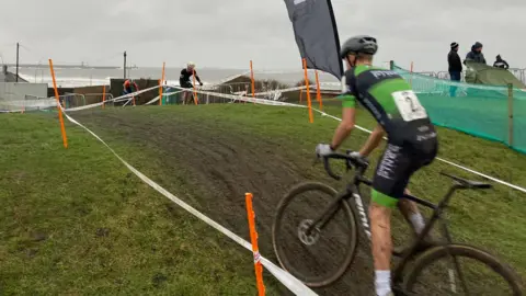 A rider in a black and green jersey has mud spattered up his legs and he is standing out of his saddle on a muddy track. Spectators are watching behind a taped boundary at each side of the track.