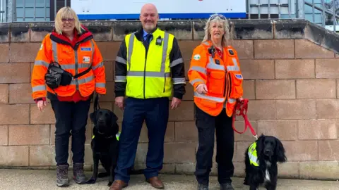 Isle of Wight Search and Rescue Catherine Fitton stands wither her black labrador Ginny, Catherine has short blonde hair with a fringe and wears glasses. She wears a bright orange high vis long sleeved jacket, with a red zip up fleece underneath and black jeans and walking boots, with a cross body black bag. In the centre stands Martin Gulliver, he is bald with a stubble beard and wears a yellow high vis jacket with long black sleeves and blue trousers and brown leather shoes. On the right, Jasmine Light wears a similar orange high vis coat to Catherine and is holding her dog Malli, a black sprocker spaniel. They are all smiling at the camera standing in front of a brick wall.