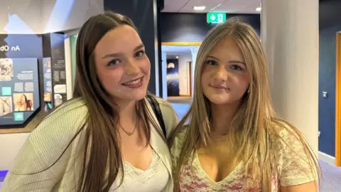 Emma and Tara are two young girls who are standing side by side. Emma on the left has long, dark hair and Tara on the right has long, blonde hair. Both girls are smiling at the camera.