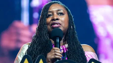 Getty Images Dawn Butler with long braided hair and carrying a black mike and behind her is a large TV screen.
